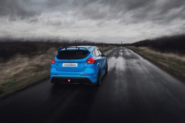 The image shows a blue Ford Focus RS MK3 dirivinf down a country road during stormy weather.